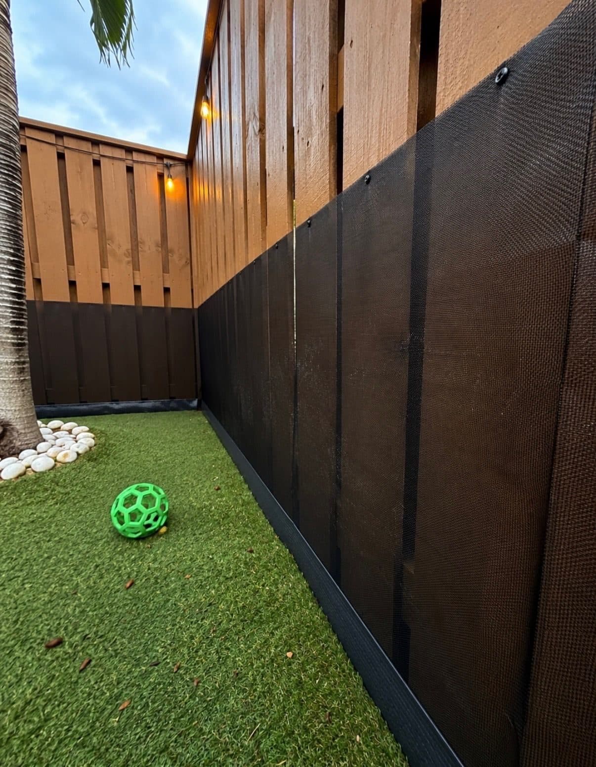Close-up of black cane toad barrier mesh installed along the base of a wood privacy fence, sealed at the ground